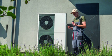 A technician in work overalls checking a heat pump unit with a tablet outside a house.