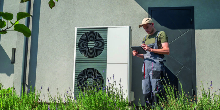 A technician in work overalls checking a heat pump unit with a tablet outside a house.