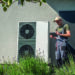 A technician in work overalls checking a heat pump unit with a tablet outside a house.